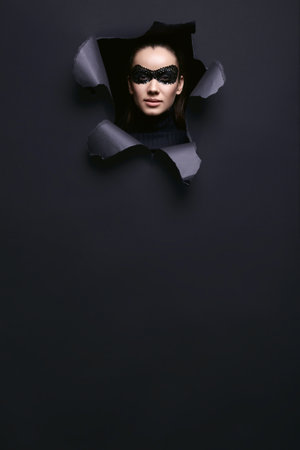 Portrait of charming elegant brunette woman in black turtleneck sweater and sequins mask looks through a ripped studio backgroundの写真素材