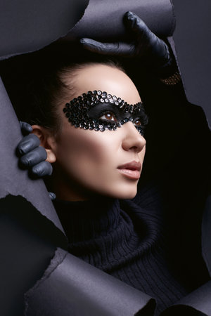Portrait of charming elegant brunette woman in black turtleneck sweater and sequins mask looks through a ripped studio backgroundの写真素材