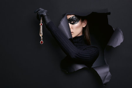 Portrait of charming elegant brunette woman in turtleneck sweater and sequins mask with black gloves posing with a stolen diamond necklaceの写真素材