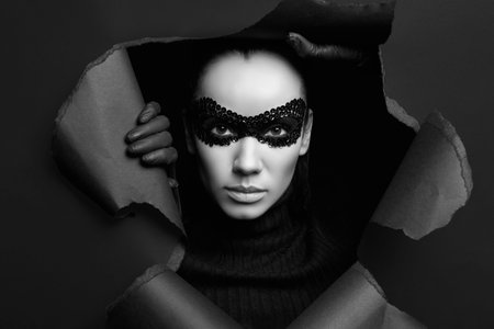 Portrait of charming elegant brunette woman in black turtleneck sweater and sequins mask looks through a ripped studio backgroundの写真素材