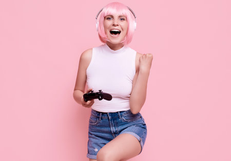 Portrait of gorgeous happy sexy gamer girl with pink hair playing video games using joystick on colorful background in studioの写真素材