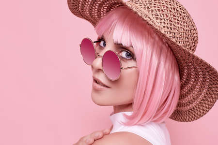 Bright summer portrait of a positive, gorgeous girl with pink hair, sunglasses and a braided hat on studio colorful backgroundの写真素材