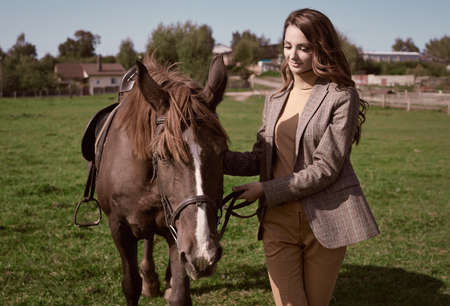 Portrait of a gorgeous brunette woman in an elegant checkered brown jacket posing with a horse on country landscapeの写真素材