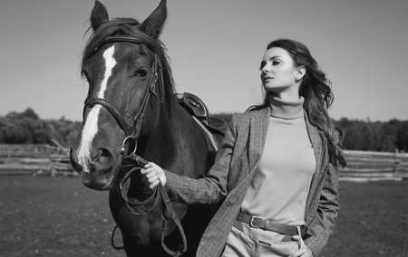 Portrait of a gorgeous brunette woman in an elegant checkered brown jacket posing with a horse on country landscapeの写真素材