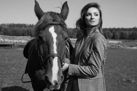 Portrait of a gorgeous brunette woman in an elegant checkered brown jacket posing with a horse on country landscapeの写真素材
