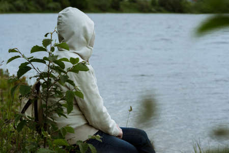 lonely woman sitting back in a jacket with a hood on the banks of the riverの写真素材