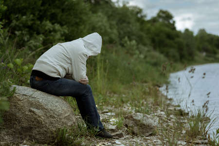 lonely woman sitting back in a jacket with a hood on the banks of the riverの写真素材
