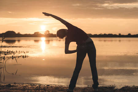 young slim woman doing exercises on the shore of the lake at sunsetの写真素材