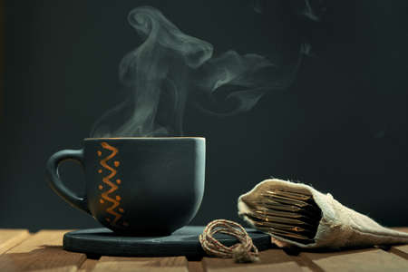 Horizontal shot of cup of hot drink with steam and a bag of tea bags on the wooden table over black background.の写真素材