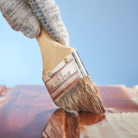 Square shot of man's hand with a paint brush painting wooden surface on a blue background.
