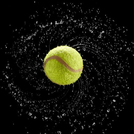 Square shot of tennis ball spinning fast splashing water drops in a circle on black background.の写真素材