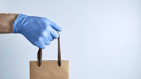 Horizontal shot of hand in a blue rubber glove holds out a paper bag on a gray background. Photo...