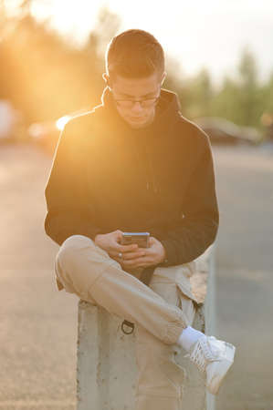 A stylish young guy in a hoodie, casual trousers and white sneakers. A guy with headphones is sitting and using a smartphone. Photo toned orange.の写真素材