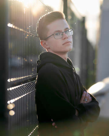 Portrait of a young stylish guy with glasses. A serious young man is standing near an iron fence. Sunlight falls from behind.の写真素材