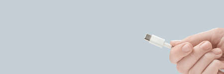 Fingers holding white type-c cable connector on gray background. Photo with copy space.