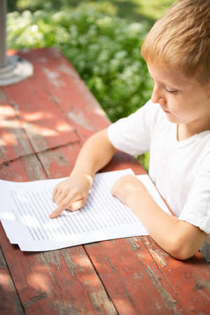 Little boy in a white T-shirt is reading outdoors. Child learns to read on the street on sunny day, preparation for school. Back to school concept.の写真素材