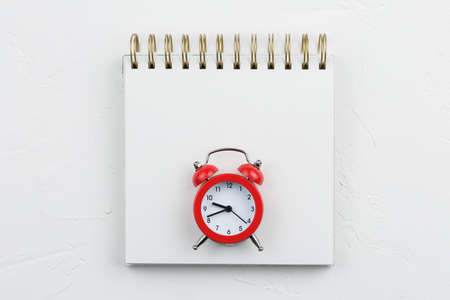 Spiral notebook with red alarm clock on white textured table. Notepad goals plans dreams, make to do list for new year concept of diary, holiday greetings or planning. Flat lay, top view, mockup.の写真素材
