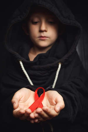 child with AID red ribbon on a dark background. the boy holds the symbol of the fight against HIV,...