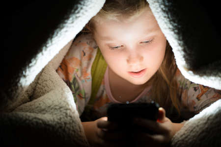 girl plays games on a smartphone at night under a blanket on the floor, the child rewrites with...