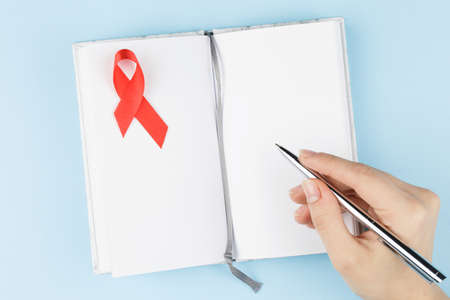 red aids ribbon on open notebook with hadns isolated on blue background, aids day and symbol of helping cancer patientsの写真素材