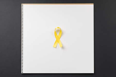 yellow cancer ribbon on notebook lying on dark table top view, gold tape world day to fight cancer...