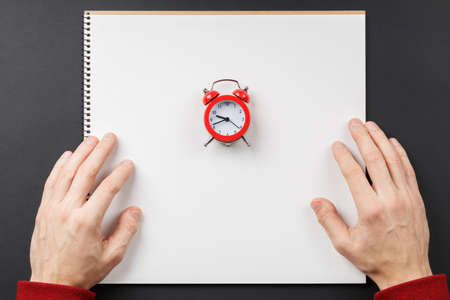 open notebook with red alarm clock in hands on black background top view, school notepad on a dark...