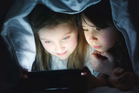 two girlfriends play on the phone under the covers, two little girls watch cartoons on the...