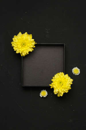 Black empty box packaging. Top view with yellow chrysanthemum and white chamomile flowers. Flat lay, spring background At blank copy space, over black background. Vertical photo.の写真素材
