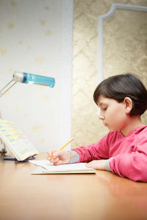 Little Asian schoolgirl doing homework while sitting at the desk at home. The girl writes with a...