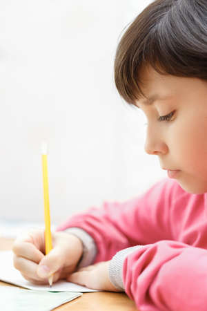 Little Asian schoolgirl doing homework while sitting at the desk at home. The girl writes with a pencil in a notebook. In the evening. Vertical image.の写真素材