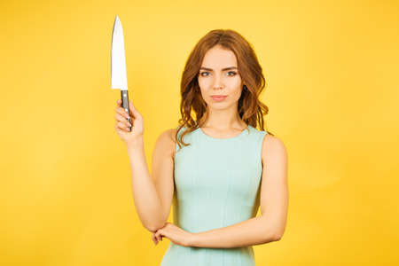 Beautiful woman in a cyan dress with a large kitchen knife in her hands on a yellow background....
