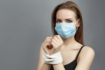 Woman with tied hands in a medical mask on a gray background studio photo Violent action concept Human traffickingの写真素材