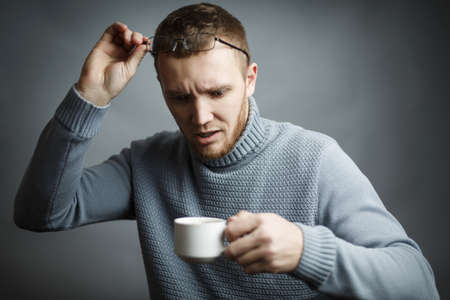 Man in sweater raised his glasses in surprise looks at a cup of espresso in his hands on a gray...