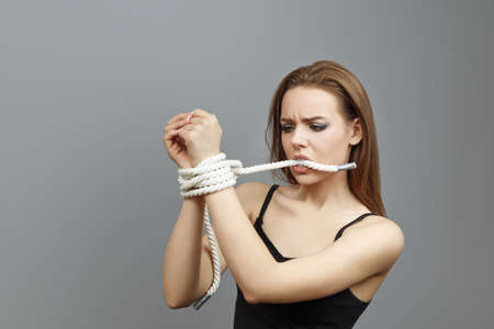 Woman with tied hands trying to untie the rope with her teeth on gray background studio photo...