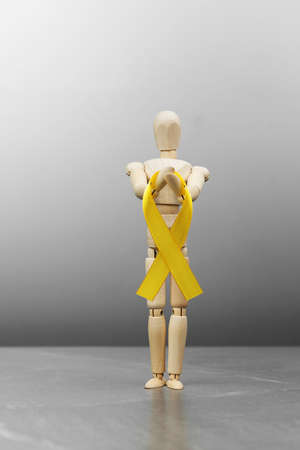 Wooden mannequin doll holds gold Childhood cancer ribbon on gray background. Cancer Control Concept. Symbolizing the day of AID, bladder, sarcoma and bone cancer.の写真素材