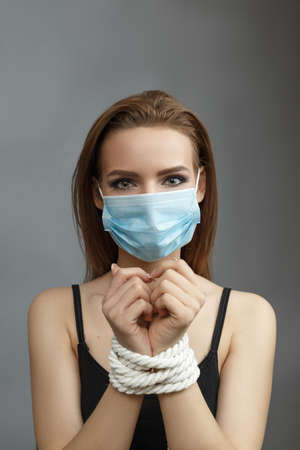 Woman with tied hands in a medical mask on a gray background studio photo Violent action concept...