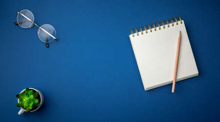 Spiral notebook with a pencil, glasses and green plant in a cup on blue background. Office table top view concept.  Close up photo.の写真素材