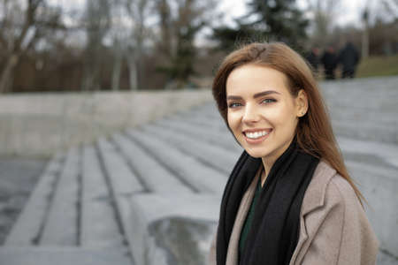 Beautiful woman in a gray coat spending time outdoors. Resting on a granite bench. She smiles and is...