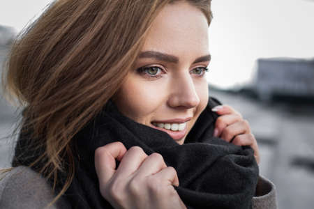 Beautiful woman wraps herself in a scarf in a gray coat on the street near concrete benches....