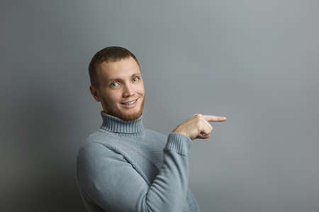 Smiling guy is pointing with his index finger towards something. On isolated gray background studio...