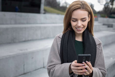 Beautiful woman stands on the street  watching on something in the smartphone, smiling and it brings...