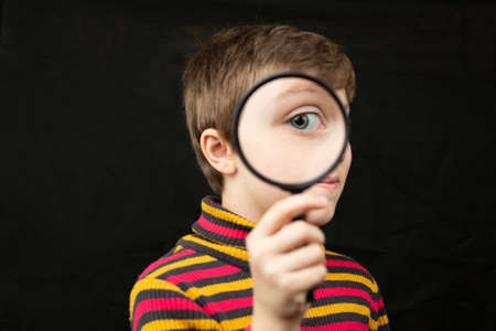 Little short haircut girl looks through a magnifier into the screen. Studio photo on a black isolated background.の写真素材