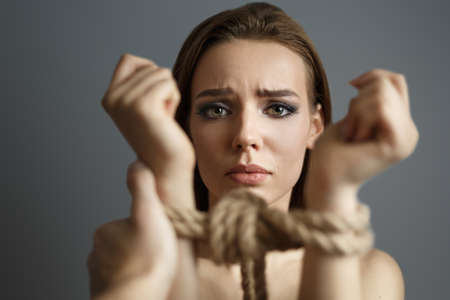 Woman with tied hands with a rope and man hand grabs her on gray background. Studio photo. Concept of violent action Human trafficking Kidnappingの写真素材