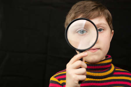 Little short haircut girl looks through a magnifier into the screen. Studio photo on a black isolated background.の写真素材
