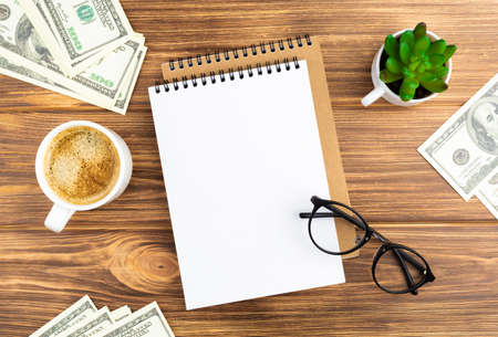 Spiral notebook with money cash doolars plant in the pot, glasses and coffee cup on wooden background. Design concept with copy space for notes. Blank page for business text. Office notepad. Top view.の写真素材