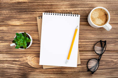 Spiral notebook with pencil, plant in the pot, glasses and coffee cup on a wooden background. Design...