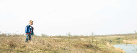 Little boy near the river canal in the countryside with last years yellow grass along the banks. The beginning of spring. Horizontal panoramic photo.の写真素材