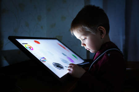 Little boy draw on a graphic tablet in the evening in a dark room under the light of the monitor....