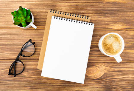 Spiral notebook with a plant in the pot, glasses and coffee cup on a wooden background. Design concept with copy space for notes. Blank page for business text. Office notepad. Top view.の写真素材