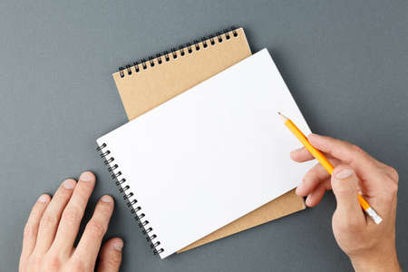 Open spiral notebook with clear page and male hands with yellow pencil on grey background. Design...